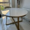 Aurora Gold Round Table with a sleek white top and gold metal base, positioned in a modern interior setting, emphasizing luxury and style for contemporary home decor.