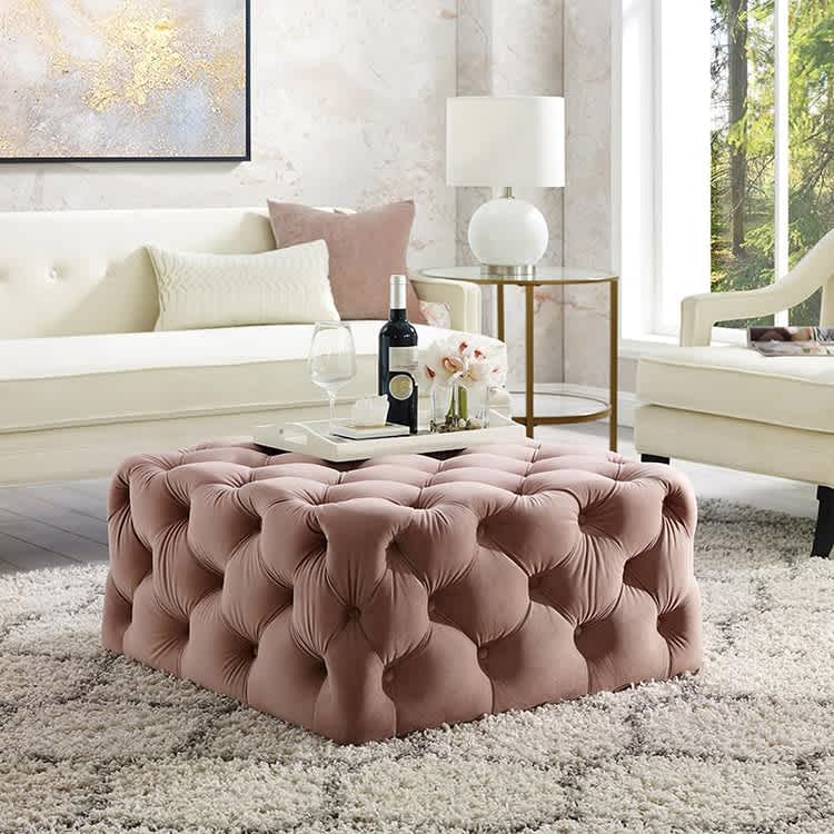 A pink velvet button-tufted ottoman functions as an elegant coffee table in a modern living room setting