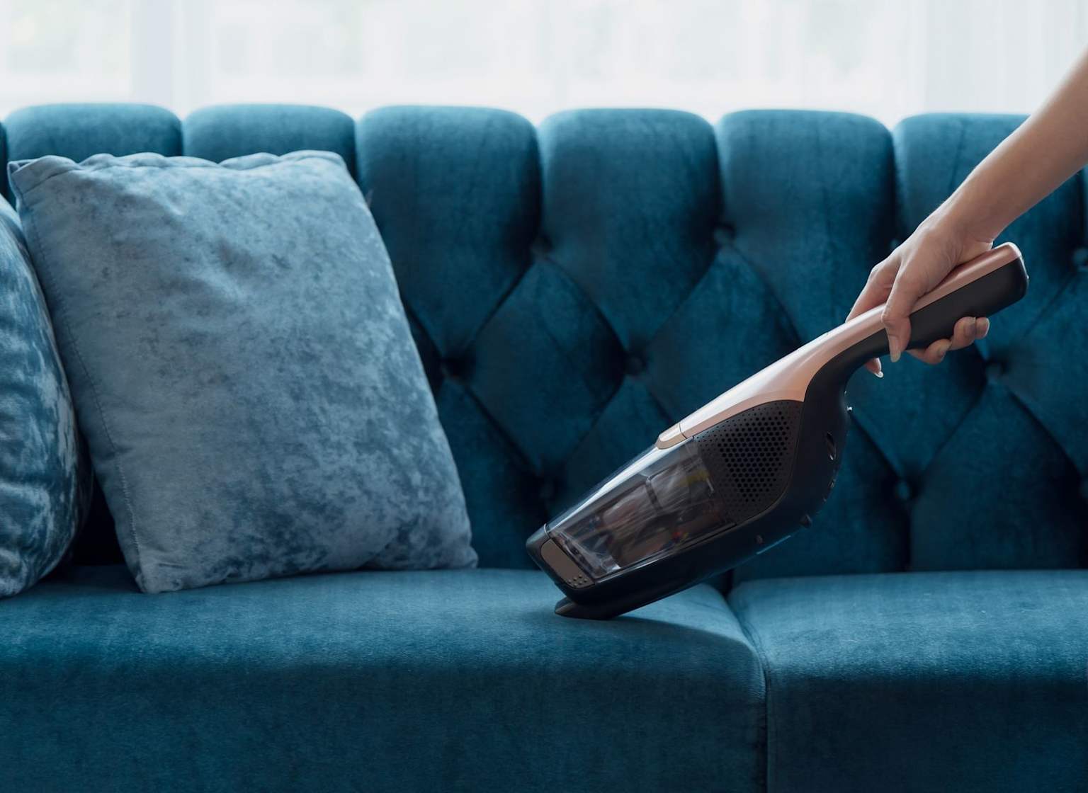 Using a handheld vacuum cleaner to clean a blue velvet sofa as part of the upholstery maintenance process