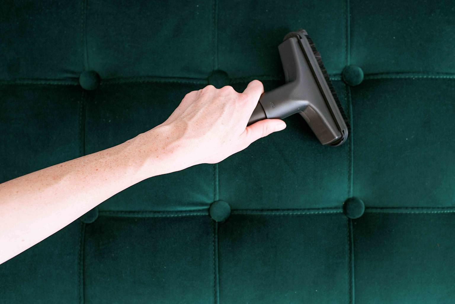 Vacuuming a dark green velvet sofa with a specialized attachment to clean upholstery gently
